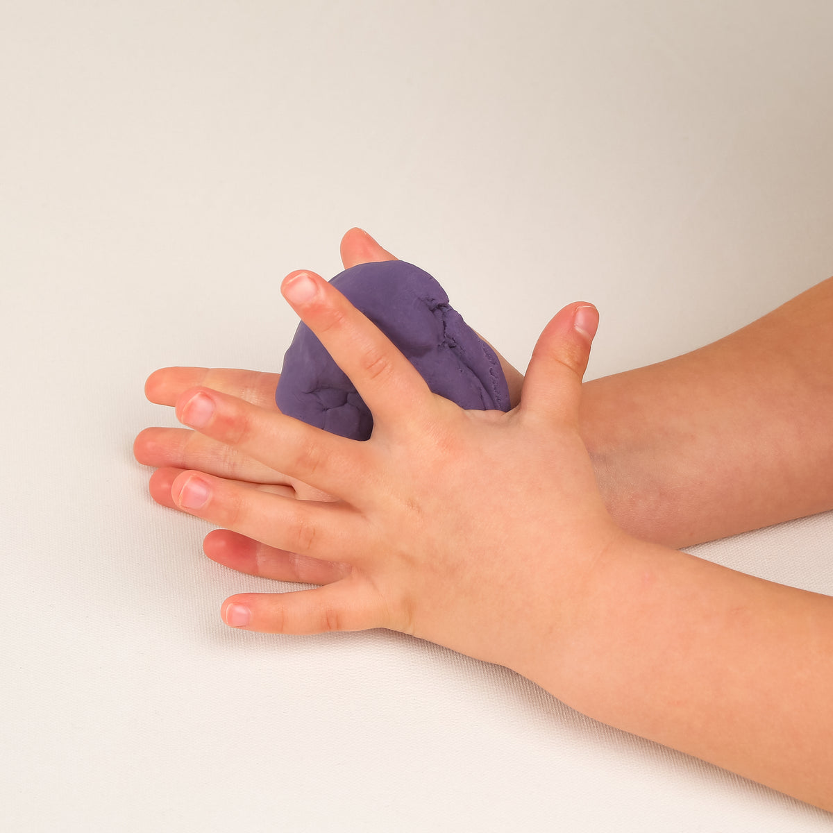 A close-up of a child's hands and forearms pressing and shaping a purple clay dough on a plain off-white background.